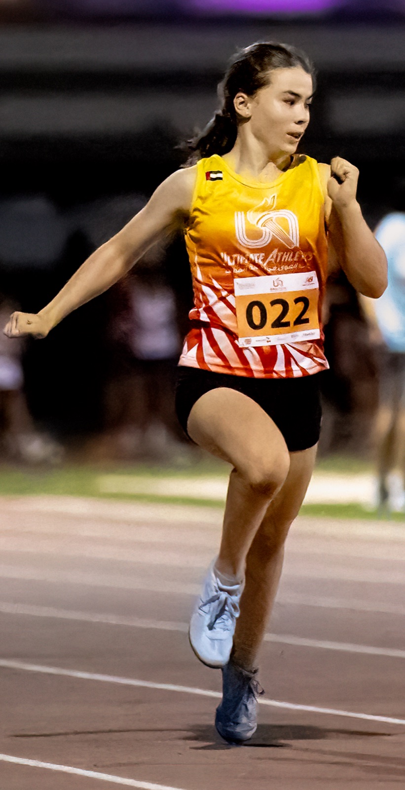 Elena running on a track at night in a yellow Ultimate Athletics vest, bib 022