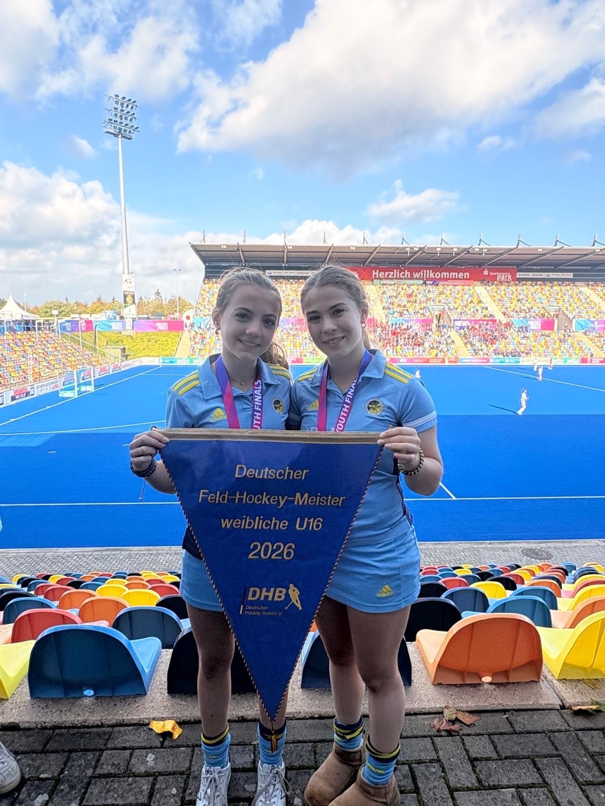 Carla and Elena holding the DHB pennant 'Deutscher Feld-Hockey-Meister weibliche U16 2026' in the stadium